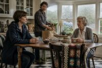 Three family members in a rain-lit Connecticut kitchen—the daughter anxious, the son quietly supportive, and the elderly mother proud yet weary—share quiet tension and hope as rain streaks the bay windows.