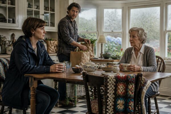 Three family members in a rain-lit Connecticut kitchen—the daughter anxious, the son quietly supportive, and the elderly mother proud yet weary—share quiet tension and hope as rain streaks the bay windows.