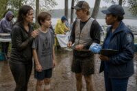Four adults and a teen stand in muddy running gear at a rainy Missouri riverfront race, exchanging uncertain, supportive looks before the charity relay.