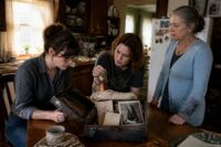 Three women—two sisters in their 30s and their older mother—gather tensely around a kitchen table, examining a scorched strongbox of old letters and keys in a sunlit, cluttered NY kitchen.
